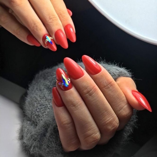 Close-up of hands with bright red almond nails, featuring iridescent rhinestone accents on the ring and index fingers.