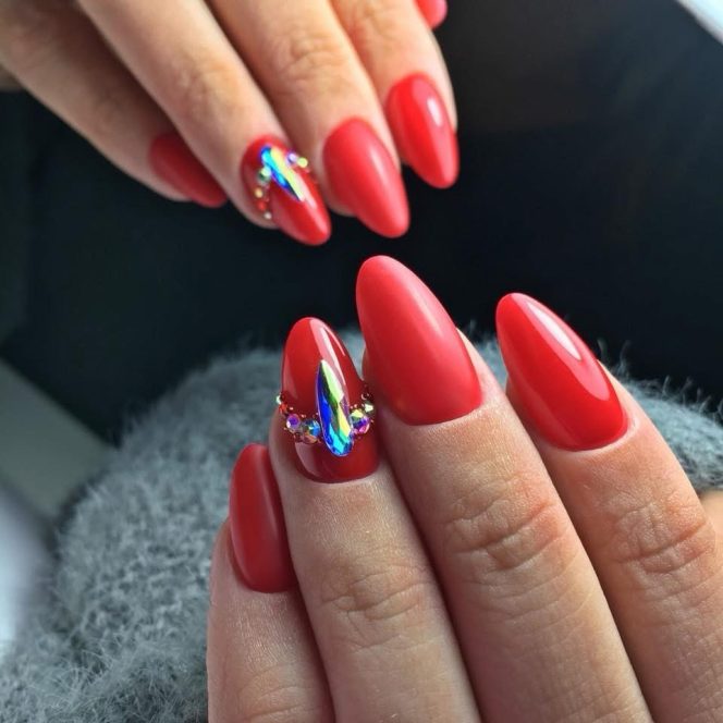 Close-up of red almond nails with a jeweled rhinestone accent on the ring finger surface of the hand.