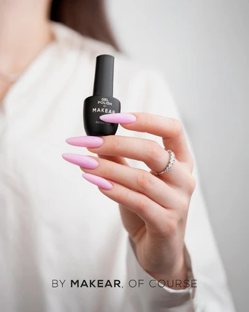 Close-up of a hand with long pink nails holding a small black bottle of Makear gel polish against a light background, with the text 'BY MAKEAR, OF COURSE' visible at the bottom.