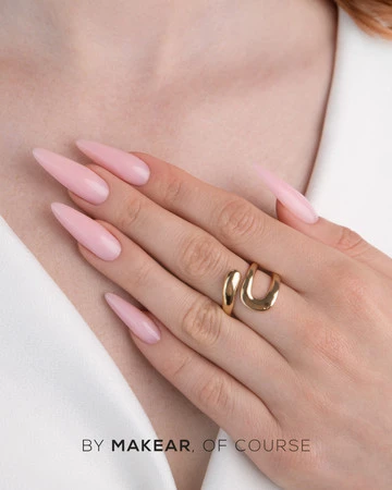 Close-up of a hand with long pink almond-shaped nails and a gold ring, with the brand text 'BY MAKEAR, OF COURSE' visible at the bottom.